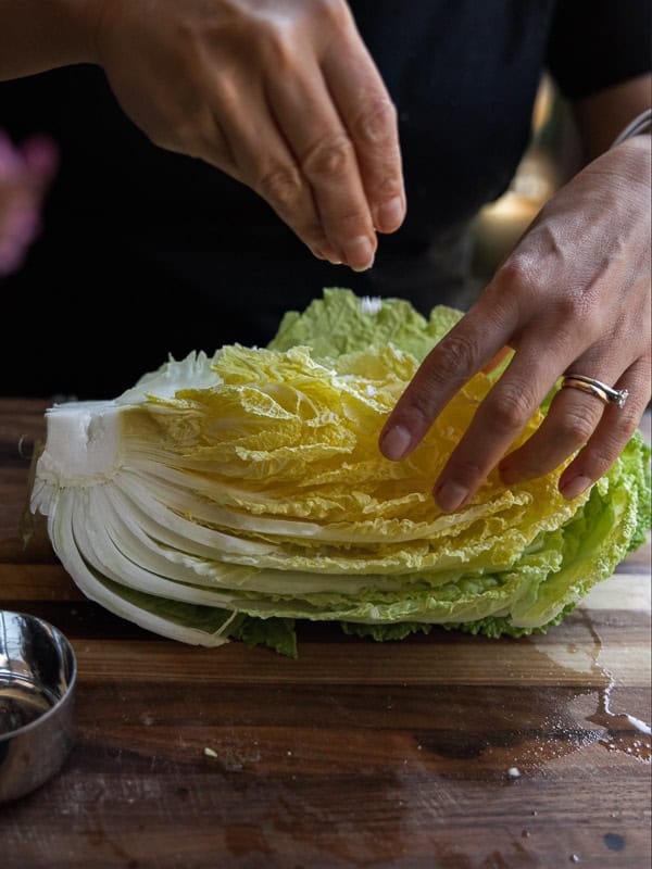 A person sprinkles salt on a halved napa cabbage on a wooden cutting board. A small metal bowl is nearby. The persons hands show a ring and are positioned over the cabbages layered leaves.